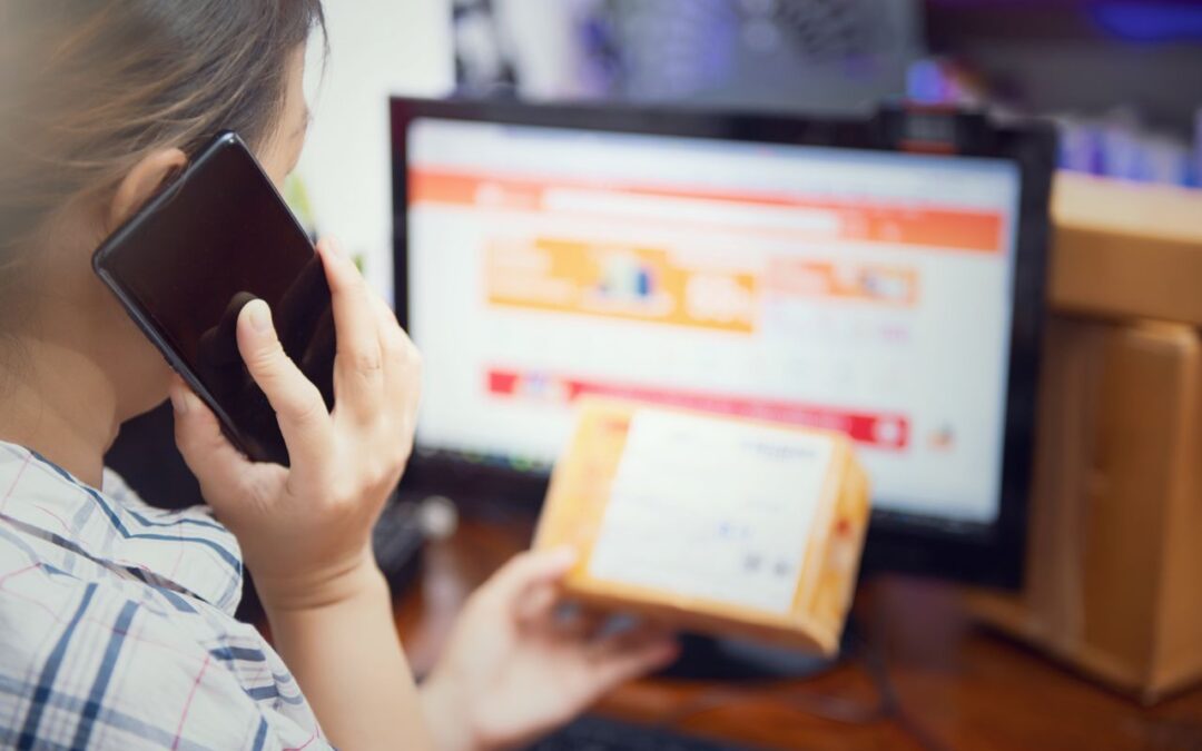 une femme effectuant un appel téléphonique tout en tenant un colis cartonné devant un écran d'ordinateur affichant un site d'e-commerce