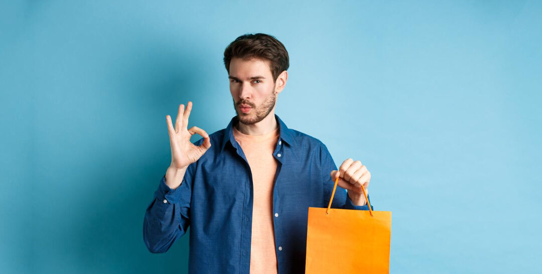 Un homme tenant un sac de shopping orange fait un signe de la main indiquant « OK » sur un fond bleu uni.