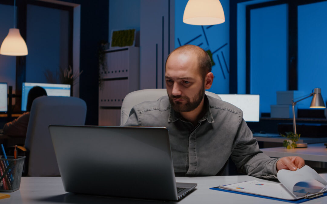 Un professionnel concentré travaille tard le soir dans un bureau en pénombre, assis face à son ordinateur portable tout en feuilletant un dossier de documents posé sur son bureau.