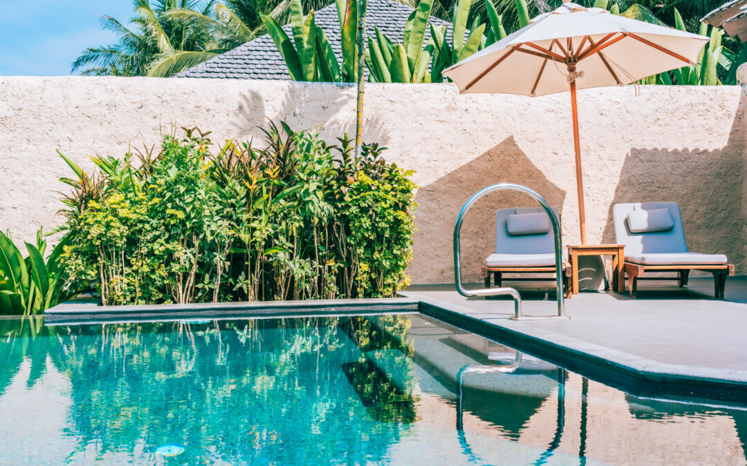 Un aménagement extérieur moderne et tropical comprend une piscine aux eaux turquoise bordée de verdure luxuriante, accompagnée d'un espace détente équipé de chaises longues et d'un parasol en toile sous le soleil.